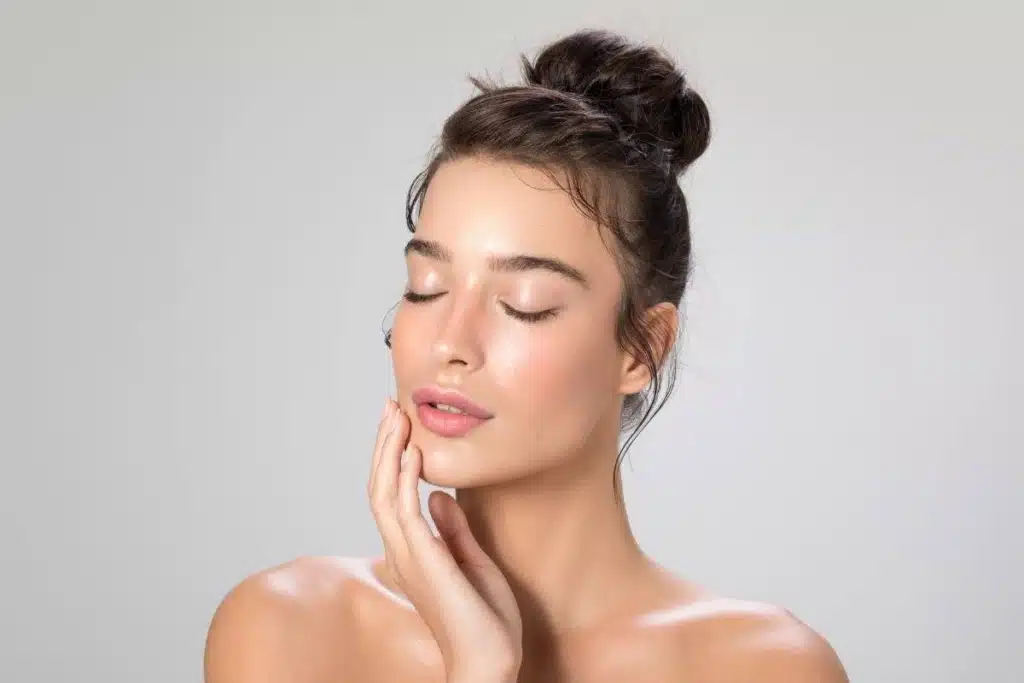 Close-up portrait of a serene woman with hair in a bun touching her glowing face with eyes closed.
