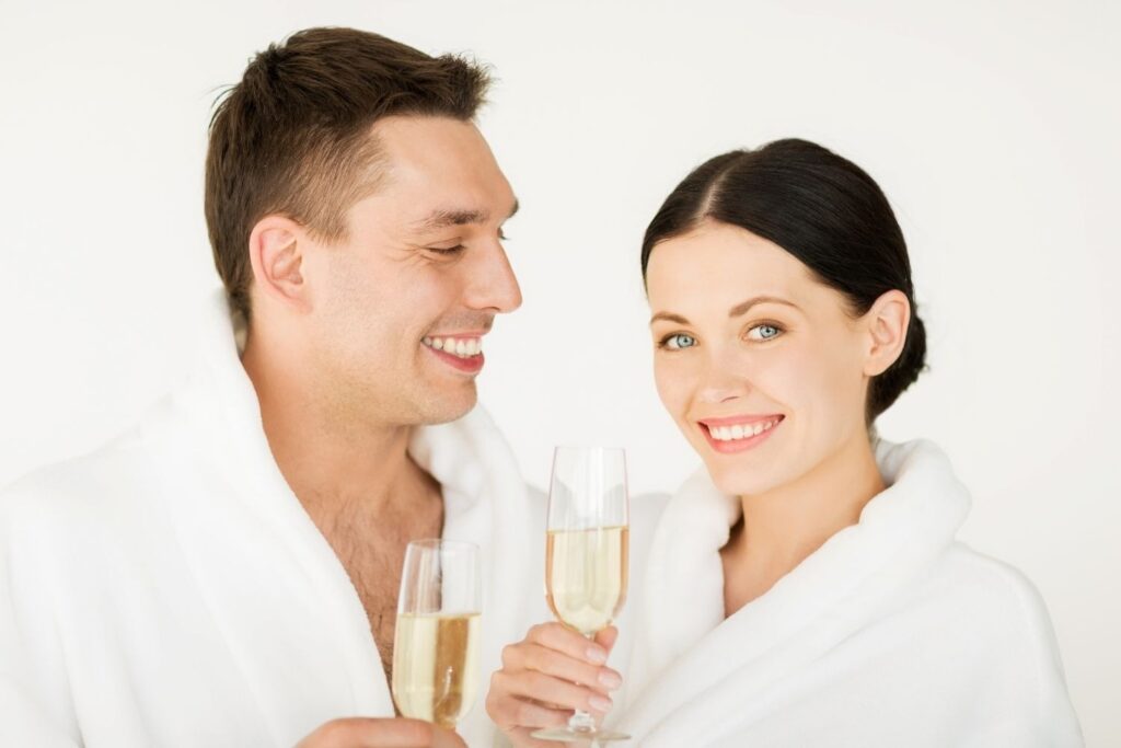 A smiling couple in white bathrobes holds champagne glasses, celebrating together and enjoying a memorable spa gifts experience.