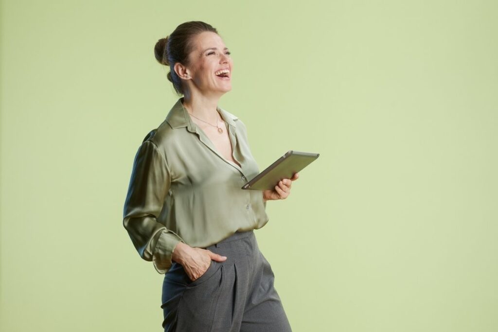 A woman in a green blouse and gray pants laughs joyfully while holding a tablet against a light green background, showcasing relaxation and IV therapy benefits.
