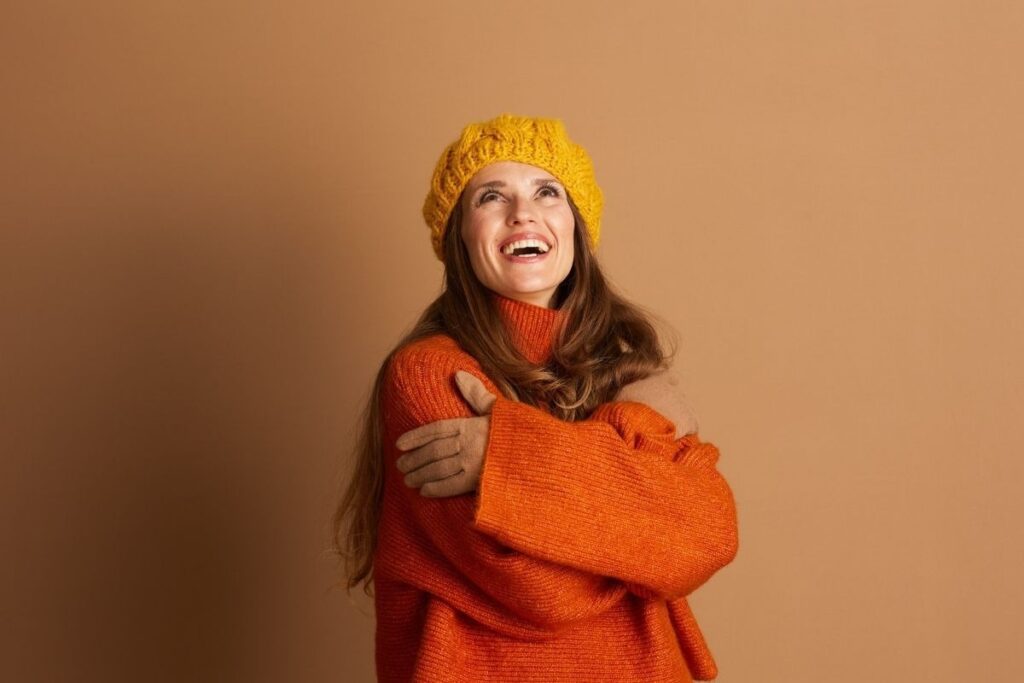 Woman in a cozy orange sweater and yellow knit hat smiles joyfully against a warm brown background, conveying comfort and the rejuvenating IV therapy benefits