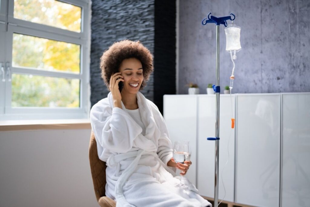 Woman holding phone and glass of water next to IV stand, following the first IV infusion guide.
