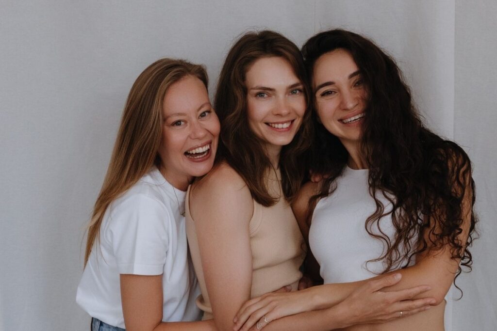 Three women stand closely together, smiling warmly against a neutral background, highlighting the truth behind IV therapy myths and wellness for active professionals.