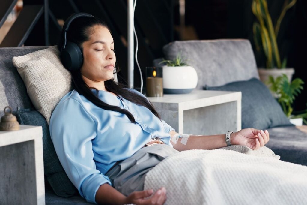 A woman wearing headphones rests on a lounge chair while receiving an IV drip treatment in a peaceful, modern setting.