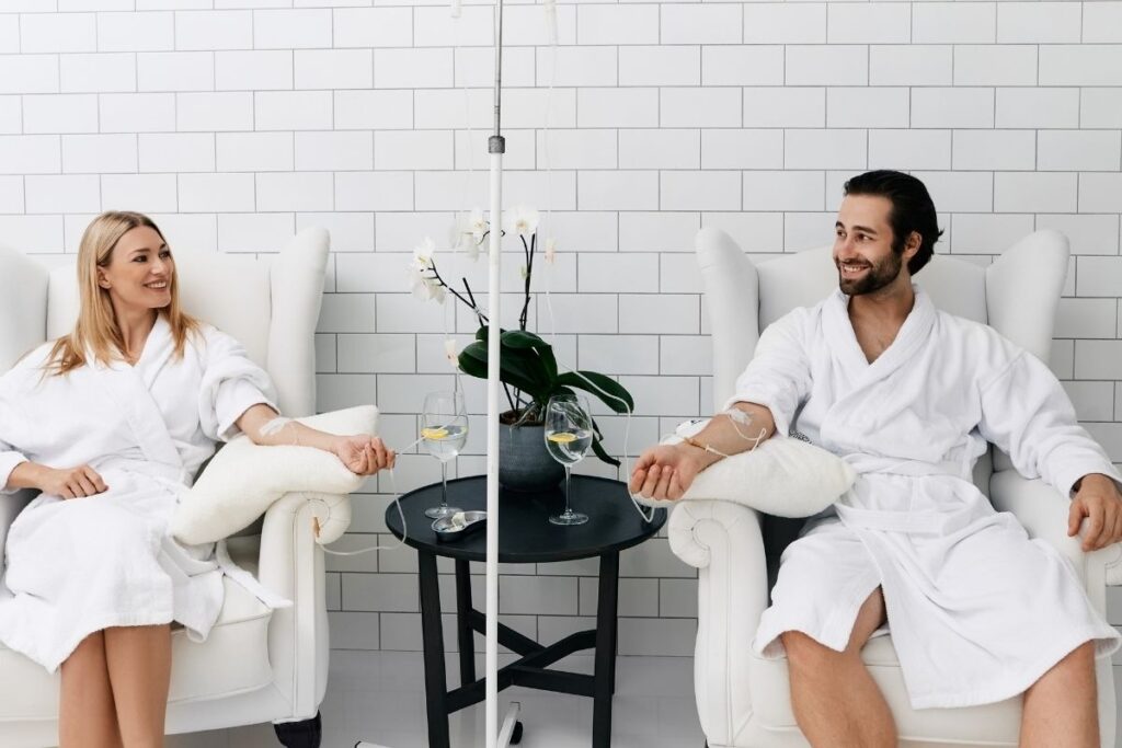 A man and woman in white robes sitting in armchairs while receiving IV drip treatments in a bright wellness clinic.