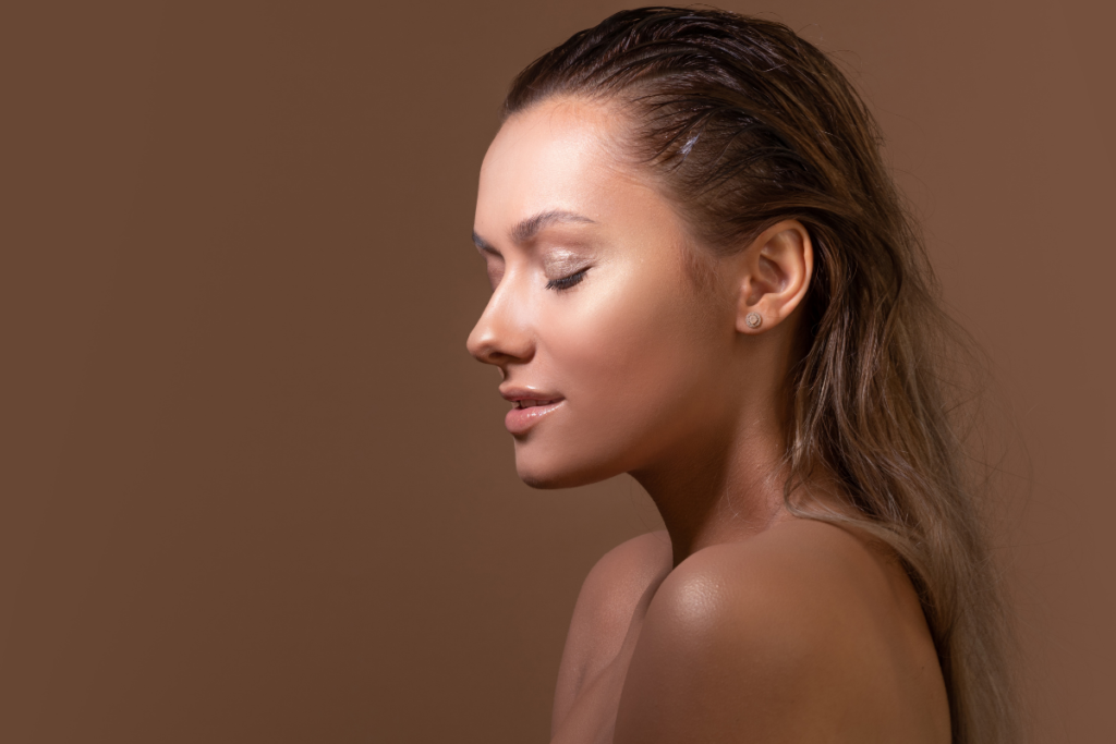 Side profile portrait of a woman with slicked-back long hair, closed eyes, and glowing bronze skin against a warm brown background
