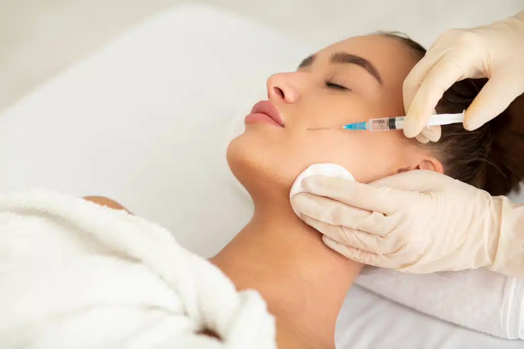 Woman lying down with eyes closed receiving a targeted facial injection from a medical practitioner wearing white gloves.