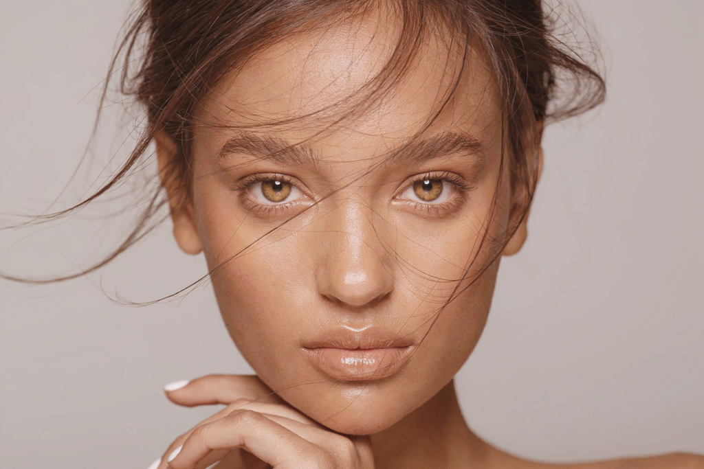 Close-up portrait of a woman with warm hazel eyes resting her chin on her hand as strands of hair sweep across her face.