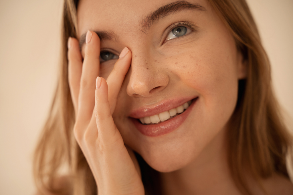 Close-up of a smiling woman with freckles gently touching her face while looking up.